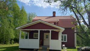 Una casa pequeña de un solo piso con tejado rojo y detalles blancos, rodeada de un césped y árboles.