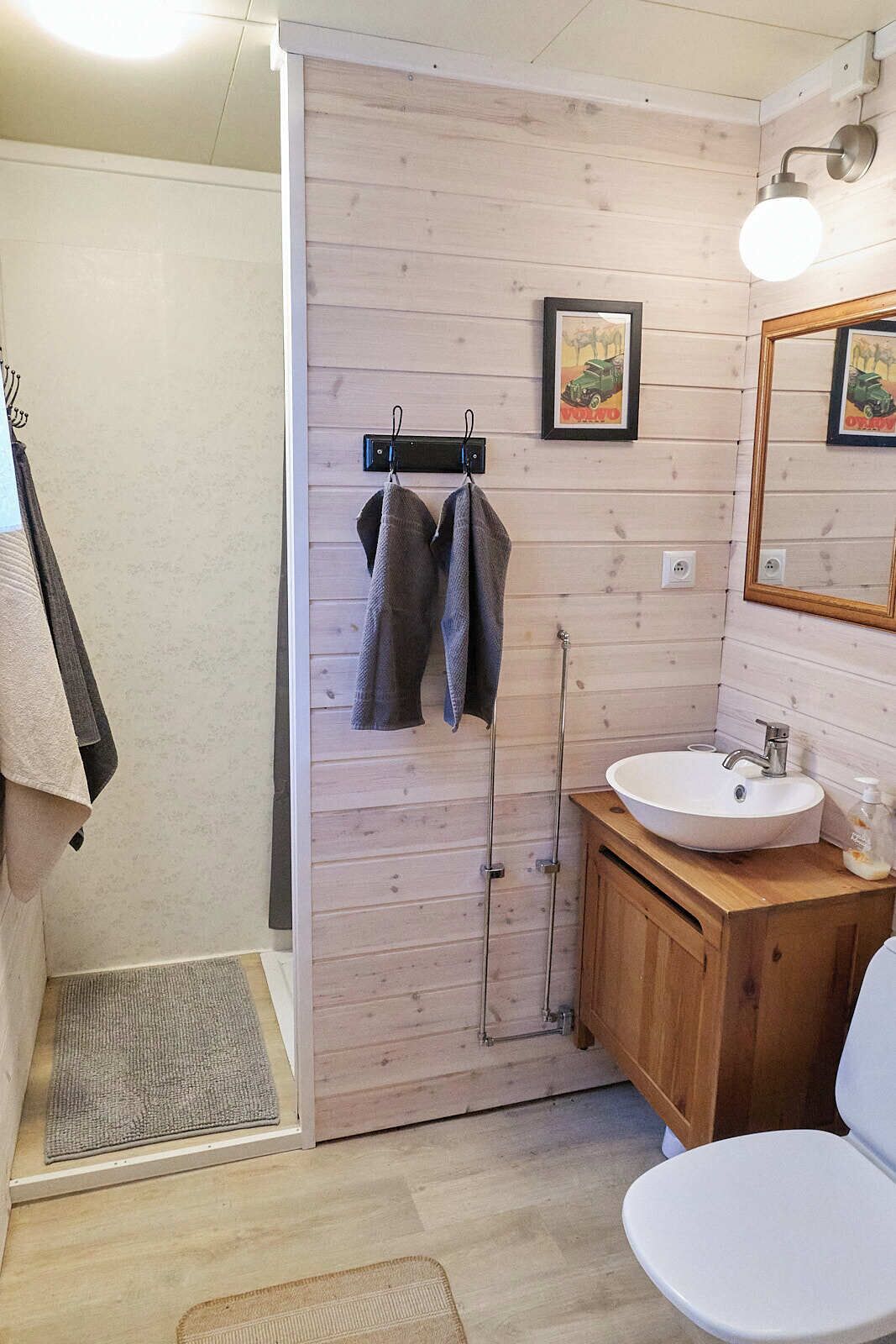 Un baño con pared de madera, un lavamanos, un inodoro y una ducha. Hay toallas colgadas en ganchos y un cuadro enmarcado en la pared.