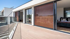 Una casa moderna con terraza de madera, grandes puertas correderas de vidrio y un dormitorio visible con cama y almohadas.