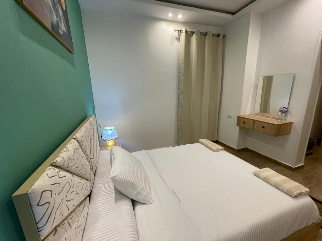 Una cama bien hecha con cabecera, una lámpara de noche, un espejo sobre el lavabo y una pared verde.