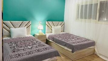 Una habitación con dos camas, una pared de acento color turquesa, una ventana con cortinas traslúcidas y una lámpara de noche.