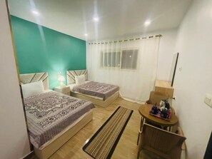 Een slaapkamer met twee bedden, een klein tafeltje, een stoel, een raam met gordijnen en een nachtkastje met een lamp.