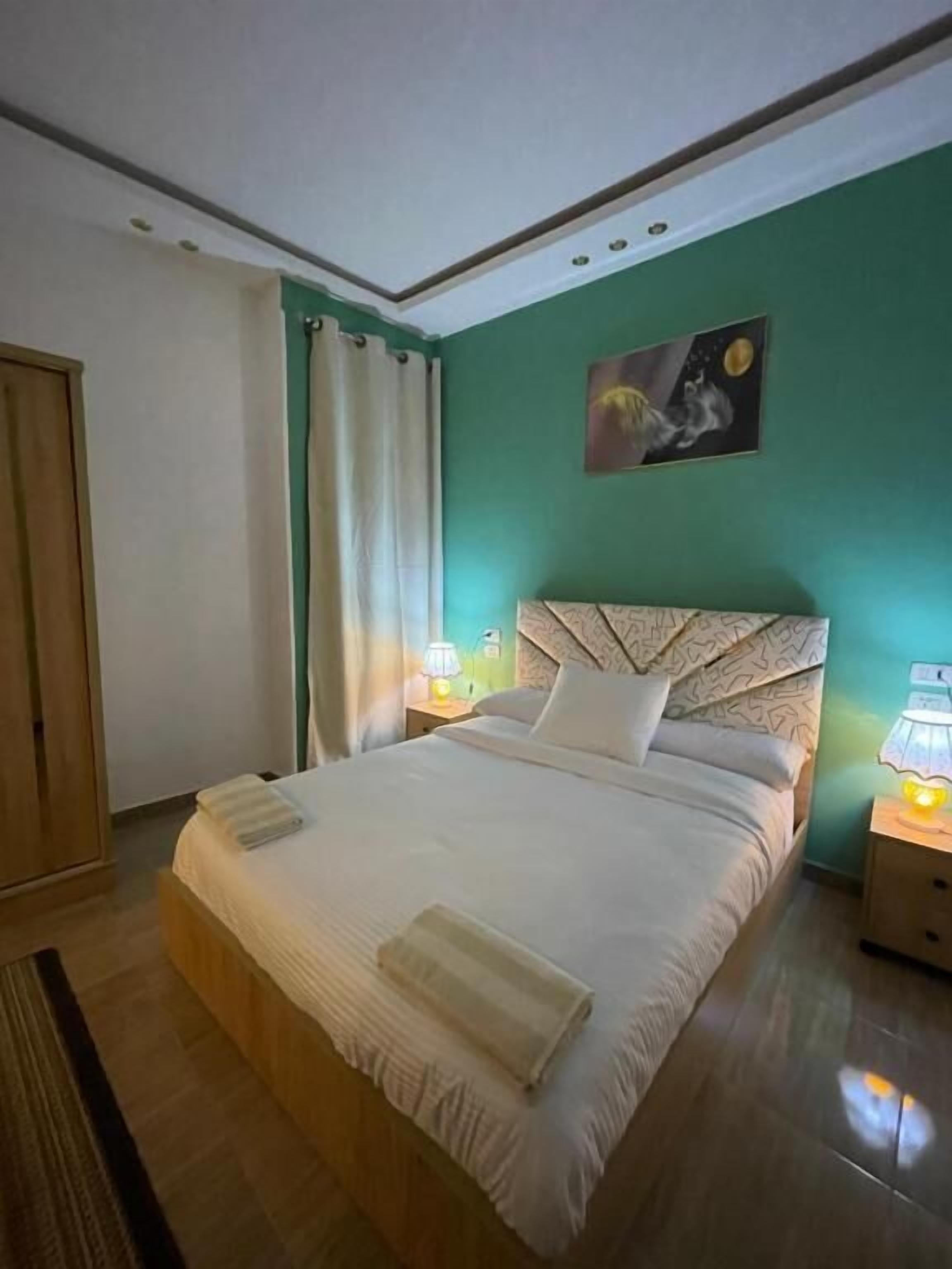 A hotel room with a bed, a nightstand, a lamp, a painting on the wall, and a door.