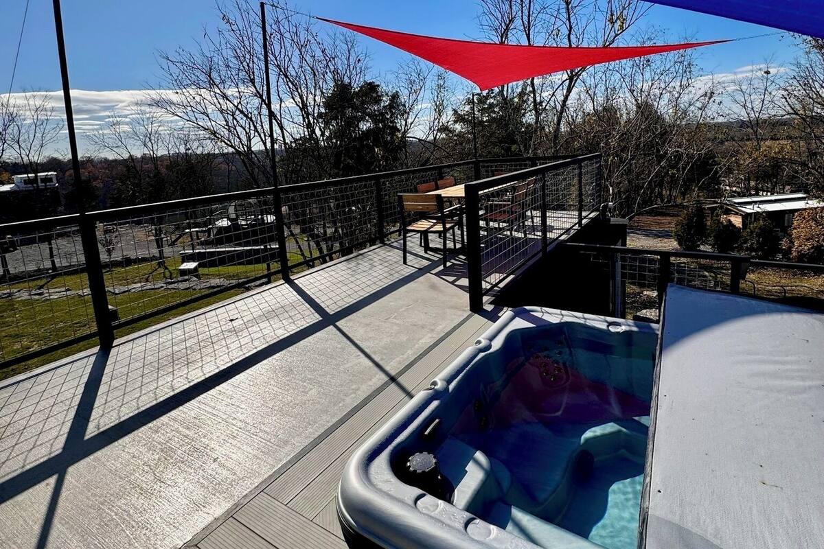 A hot tub on a patio with a blue and red shade sail overhead, surrounded by a fenced area with a view of a snowy mountain in the background.