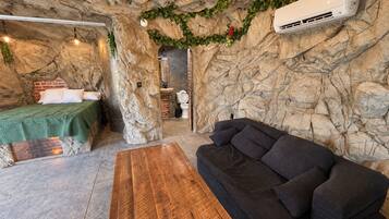 Une pièce avec un mur en pierre rustique, un lit recouvert d’une couverture verte, un canapé foncé, une table en bois et un climatiseur blanc.