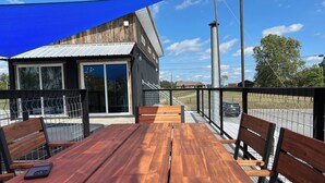 A wooden table and benches on an outdoor patio with a blue shade sail, overlooking a fenced area with a building in the background.