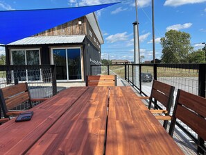 A wooden table and benches on an outdoor patio with a blue shade sail, overlooking a fenced area with a building in the background.