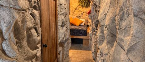 Un estrecho pasillo de paredes de piedra que conduce a una zona con camas acogedora, con una puerta de madera y una lámpara colgante.