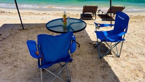 Ein Strand mit einem runden Tisch, zwei blauen Klappstühlen, einer Flasche und einem Glas auf dem Tisch.