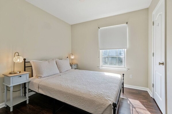 A bedroom with a bed, two bedside tables, a window with blinds, and a door.