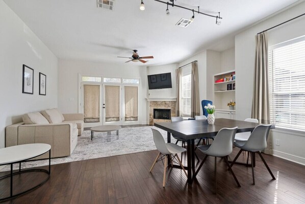 A modern living room with a sofa, a dining table with chairs, a fireplace, and a TV mounted on the wall.