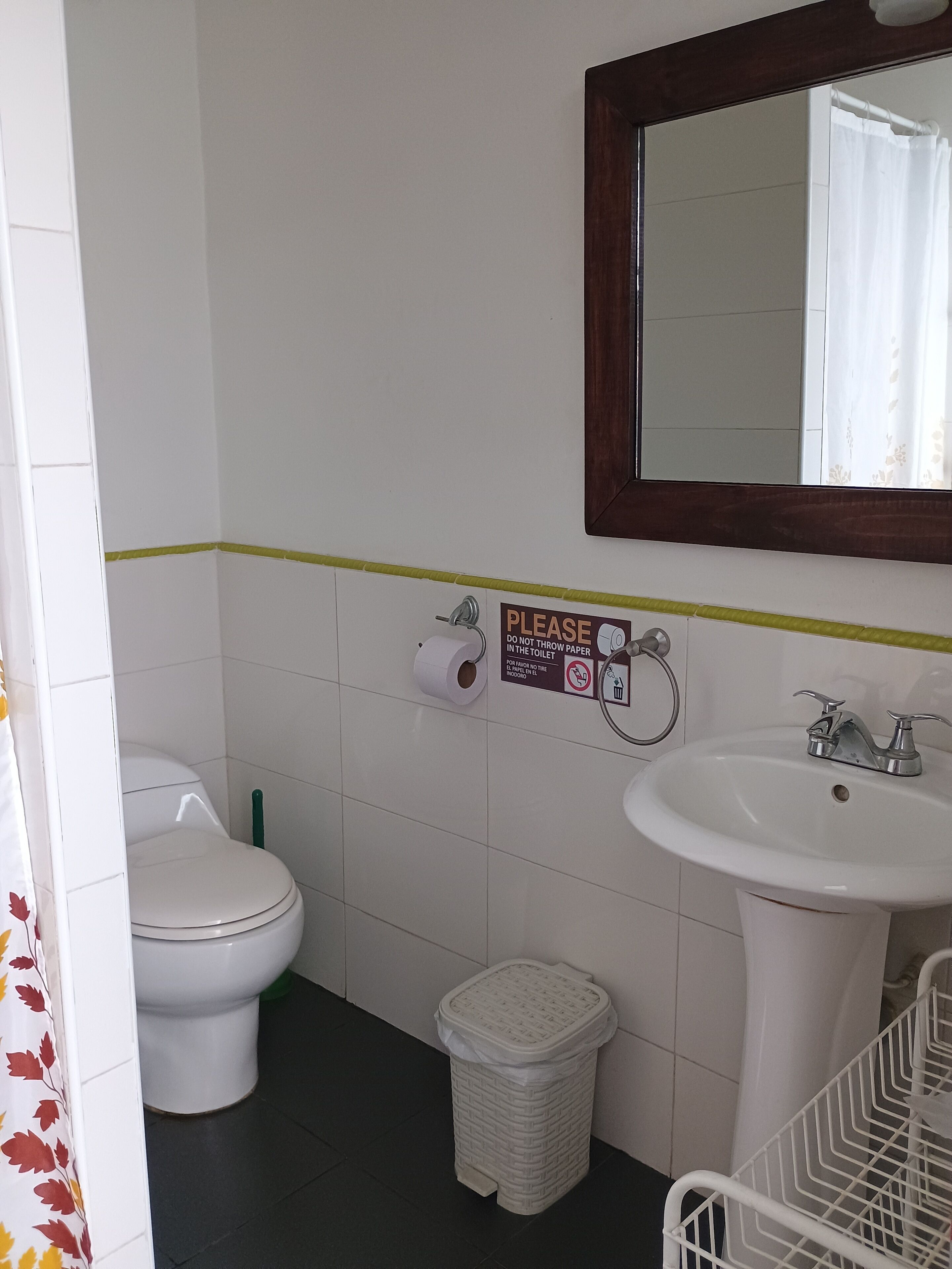 Un baño con inodoro, lavamanos y un cubo de basura. Un letrero en la pared indica no tirar papel en el inodoro.