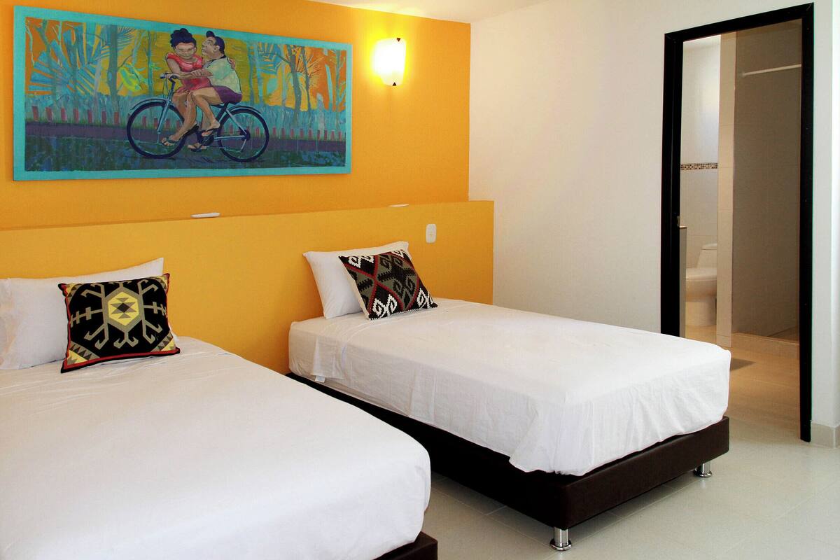 Quarto de hotel com duas camas de solteiro, um quadro na parede e um banheiro visível através de uma porta aberta.