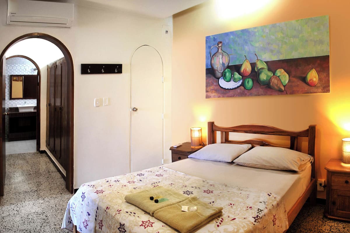 Quarto de hotel com uma cama, um quadro de frutas na parede, uma mesa de cabeceira com uma luminária e uma porta de madeira.