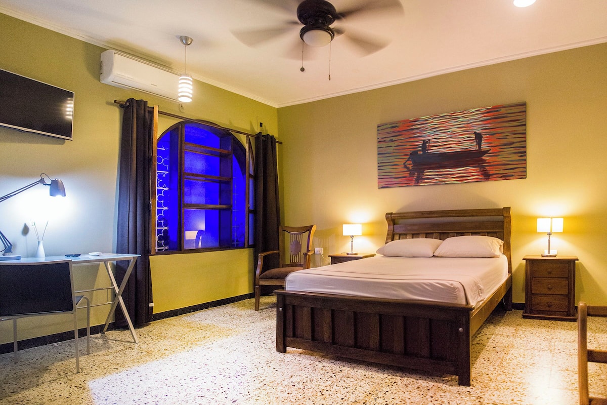 Quarto de hotel com cama, escrivaninha, cadeira e obra de arte na parede.