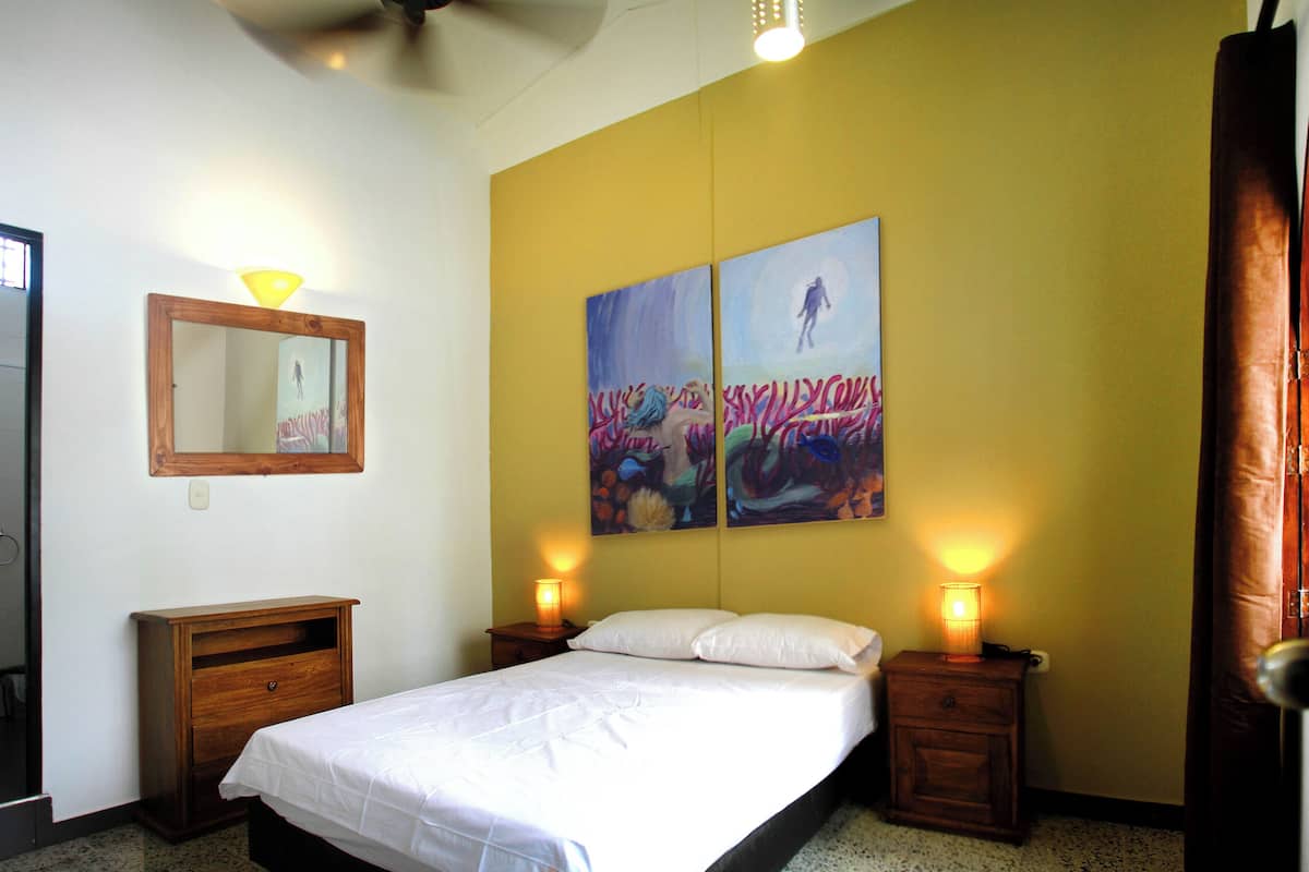 Quarto com uma cama, duas mesas de cabeceira, um criado-mudo, um espelho e duas pinturas penduradas na parede.