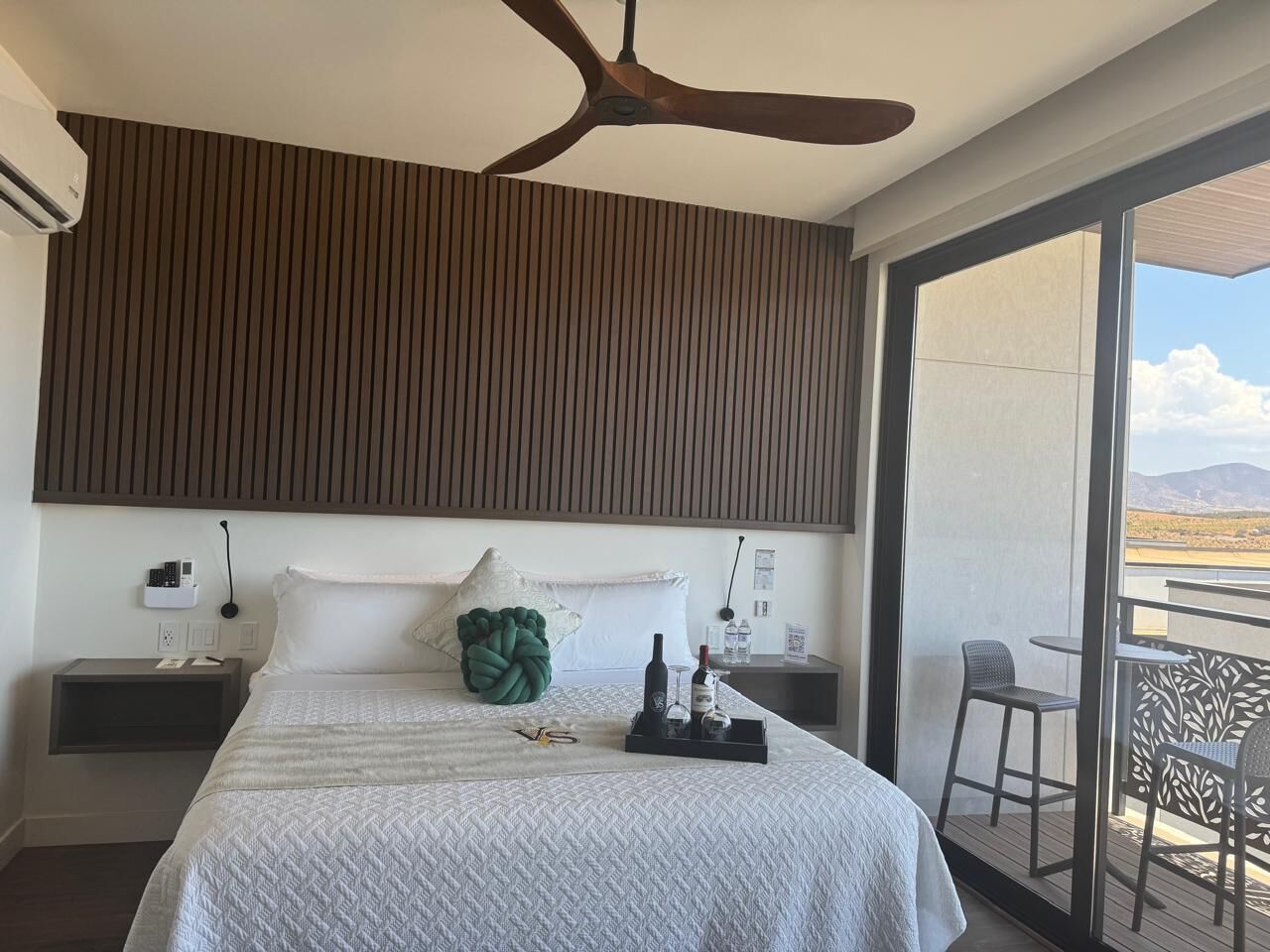 Een hotelkamer met een bed, een plafondventilator, een nachtkastje, een bijzettafeltje, een raam met uitzicht en een balkon met stoelen.