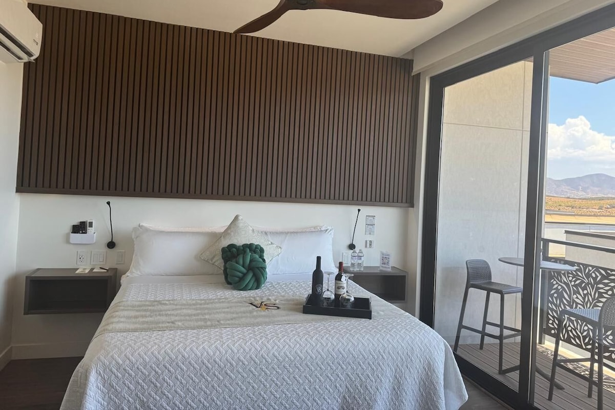 Quarto de hotel com cama, ventilador de teto, mesa de cabeceira, janela com vista e varanda com cadeiras.