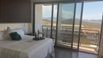 Un dormitorio con una cama grande, una mesita de noche, un cojín decorativo verde y una bandeja con objetos. La habitación tiene un amplio ventanal corredero que da a un balcón con vistas a las montañas.