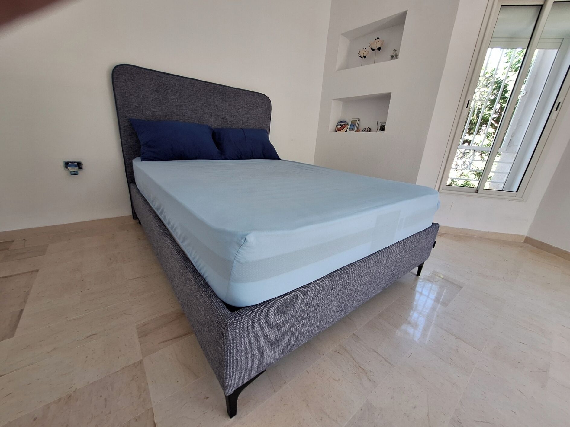 Un lit avec une tête de lit grise et une literie bleue, dans une pièce dont le sol est carrelé et qui possède une fenêtre.