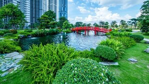 Un parque con un puente rojo sobre un estanque, rodeado de abundante vegetación y altos edificios al fondo.