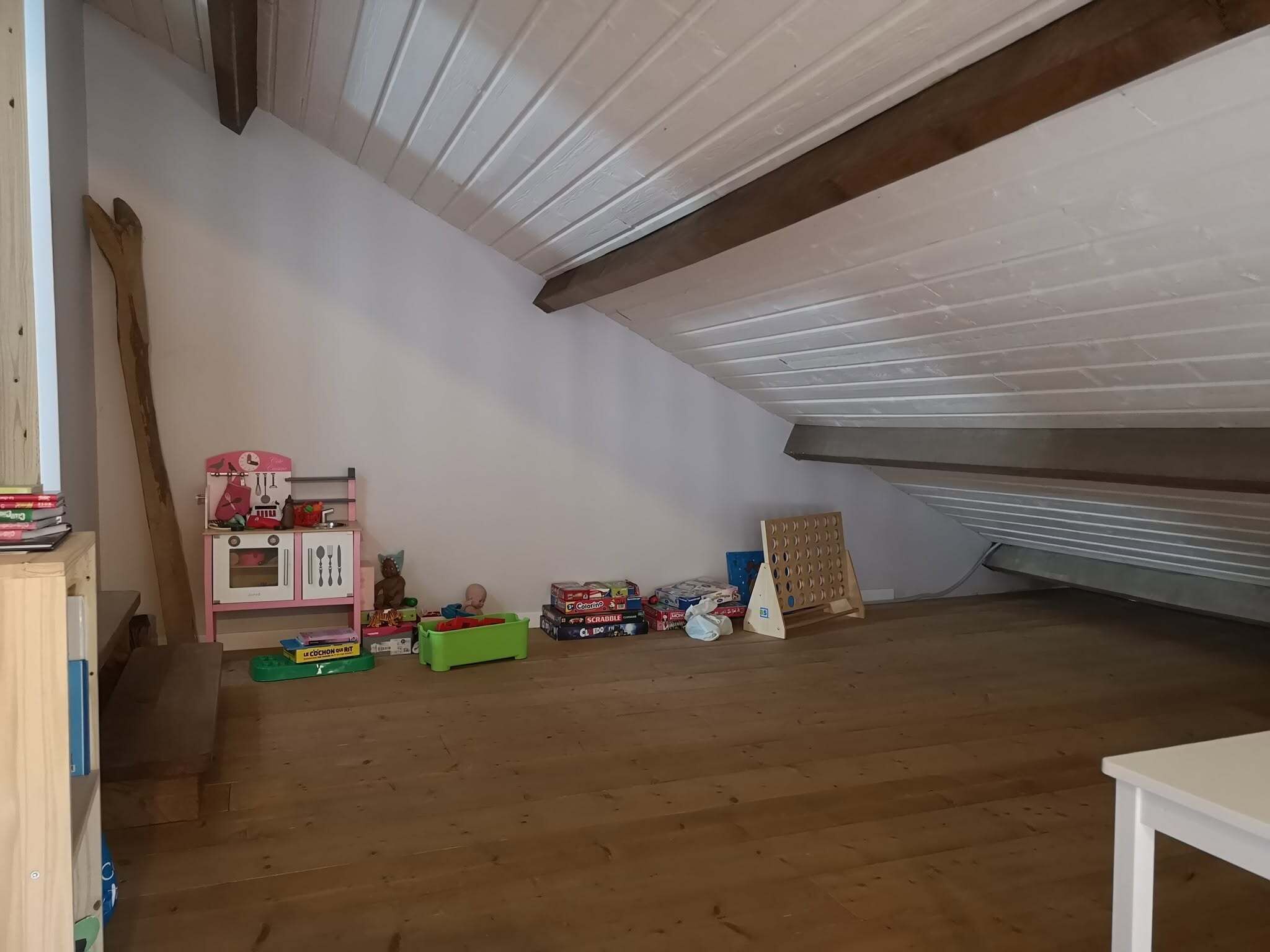 Une pièce mansardée aux hauts plafonds, dotée d’un plancher en bois, d’un petit ensemble de cuisine rose pour enfants, ainsi que de divers jouets et boîtes pour enfants.