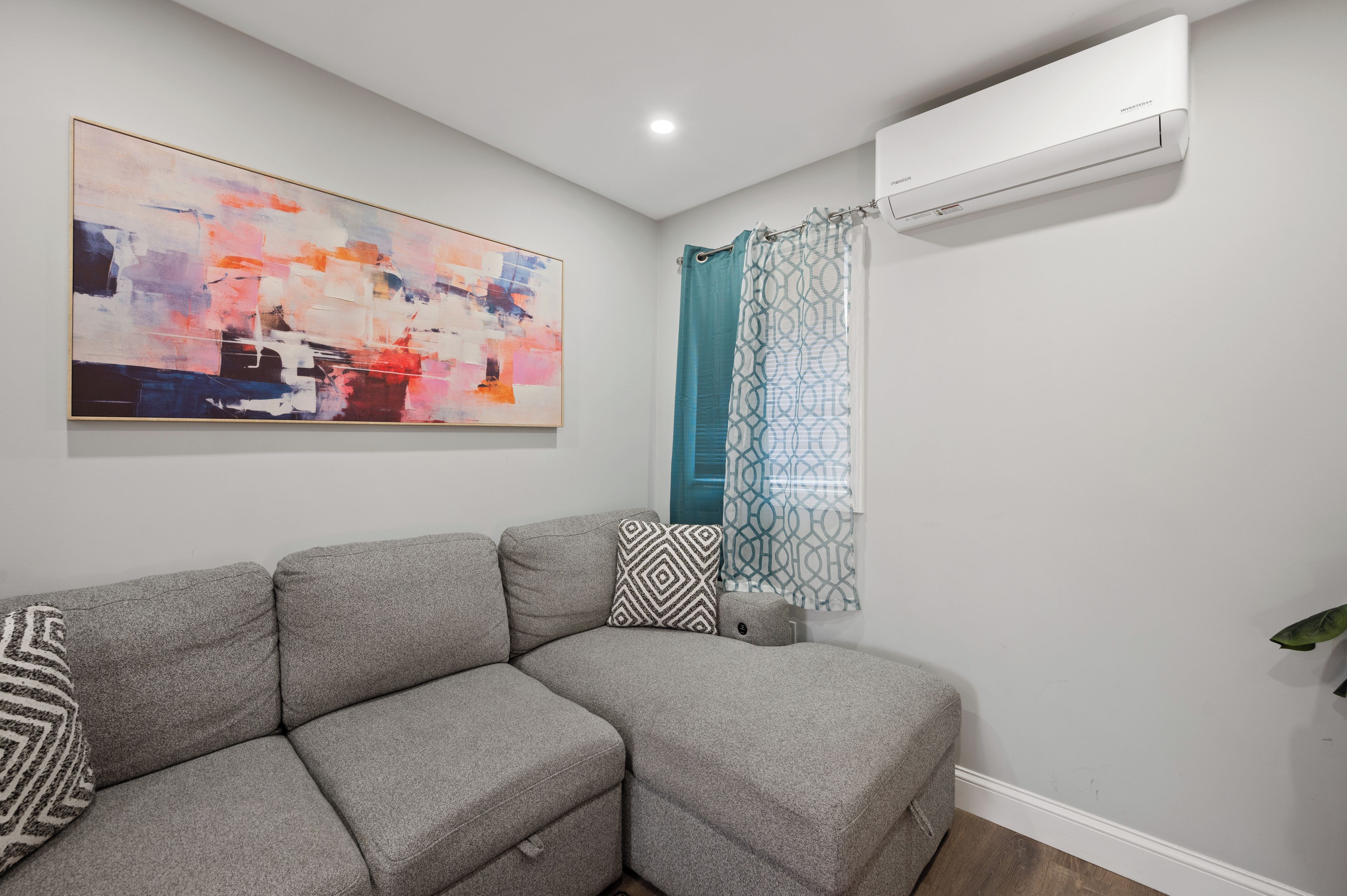 A living room with a gray sectional sofa, a large abstract painting, a patterned shower curtain, and a wall-mounted air conditioner.