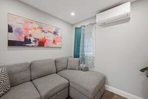 Een woonkamer met een grijze hoekbank, een groot abstract schilderij, een gordijn met een patroon voor de douche en een luchtverwarmer aan de muur.