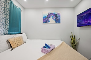 A hotel room with a bed, a painting, a television, and a plant.