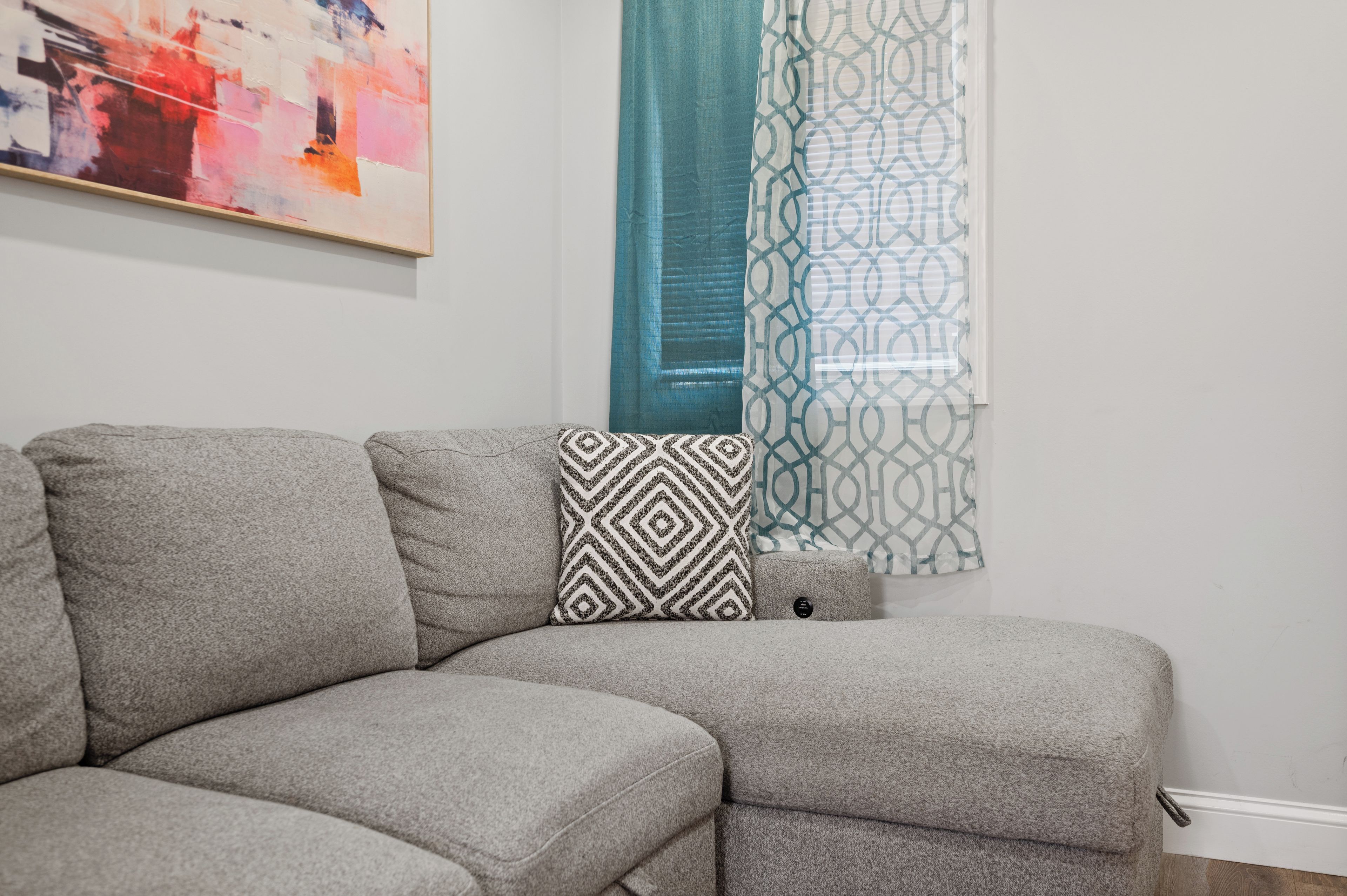 Un sofá modular con un cojín de estampado geométrico, una cortina color turquesa y una pared con un cuadro abstracto enmarcado.