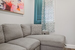 Een modulaire bank met een kussen met een geometrisch patroon, een blauwgroen gordijn en een muur met een ingelijst abstract schilderij.