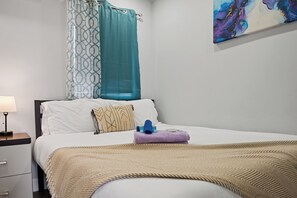 Ein Hotelzimmer mit Bett, Nachttisch, Lampe, einem Bild an der Wand und einem Fenster mit Vorhang.