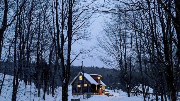 Une cabane en bois, dont le toit est recouvert de neige, entourée d’arbres sans feuilles et d’un paysage enneigé.