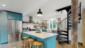 Une cuisine moderne avec des meubles de couleur turquoise, des appareils électroménagers en acier inoxydable, un îlot central doté de tabourets et un escalier agrémenté d’éléments en bois.
