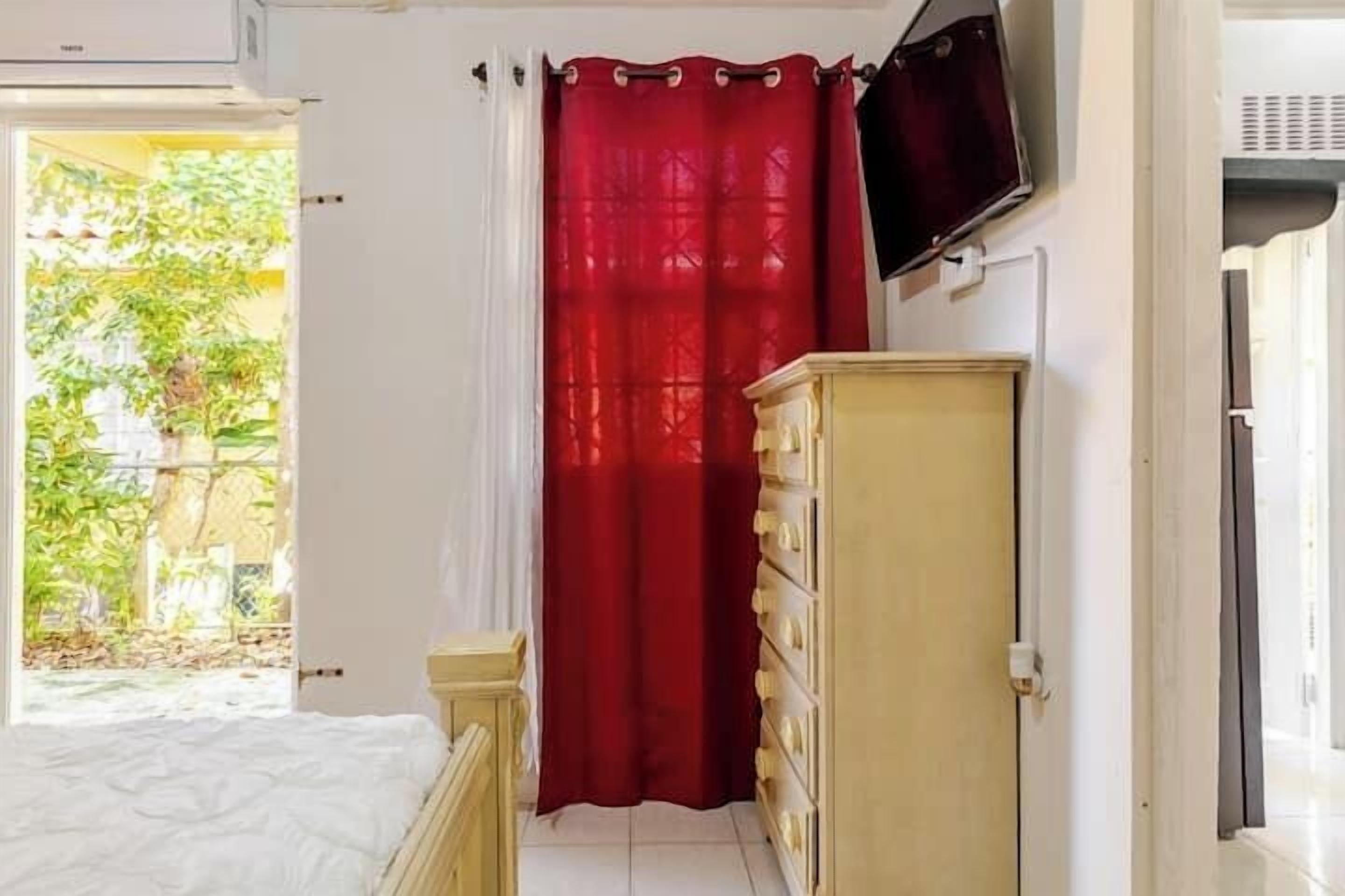 A bedroom with a bed, a dresser, a TV mounted on the wall, and a window with a red curtain.