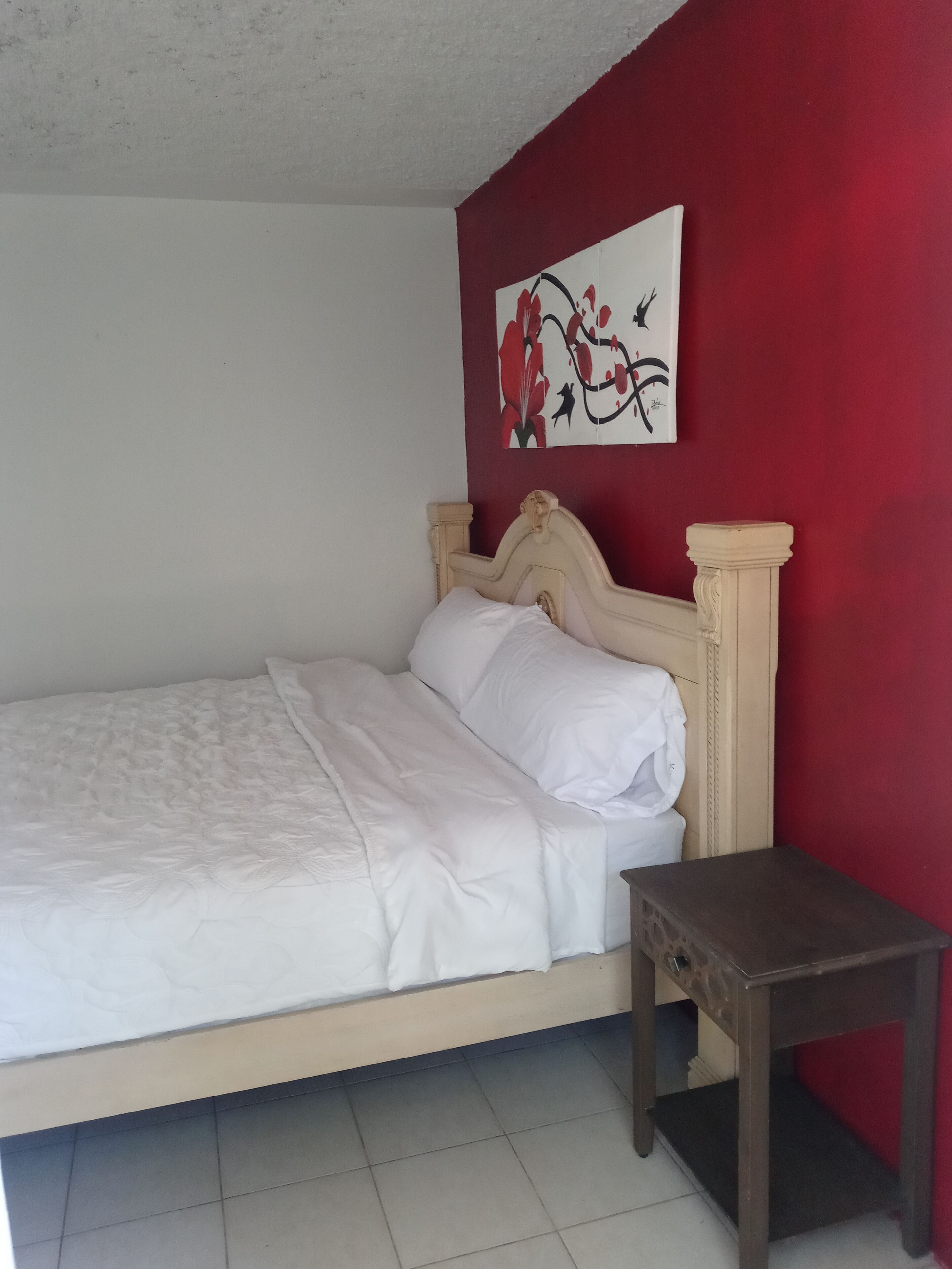A bedroom with a bed, a nightstand, and a red wall with a floral painting.