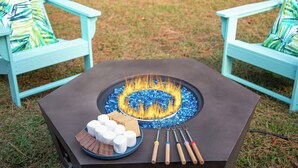 Une table carrée avec un foyer au centre, entourée de guimauves et de biscuits Graham disposés sur une assiette, ainsi qu’un ensemble de brochettes.