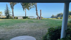 A covered patio with a round table and chair, overlooking a grassy area with trees and a view of the water. - Coastal Elements - 1BDRM - Full Kitchen (Nanoose Bay)