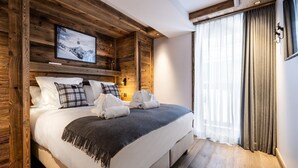 A modern bedroom with a wooden headboard, a large bed with white linens, a gray throw blanket, and a mounted television displaying a snowy mountain scene. - Wifi, 4th floor, view mountain, sauna, hammam, balcony, fireplace or stove, tv, 148m², Méribel (Méribel - Les Allues)