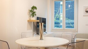 Un espace repas moderne avec une table ronde blanche, des chaises transparentes, un vase en verre et une plante en pot sur une étagère en bois.