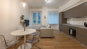 Une cuisine moderne avec un plancher en bois, une table à manger blanche et des chaises transparentes, ainsi qu’un canapé gris.
