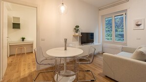 Un salon moderne avec une table ronde blanche, des chaises transparentes, un canapé beige, un téléviseur et une fenêtre donnant sur des arbres.