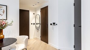 A modern hotel room with a wooden floor, a round table with a vase of flowers, a white armchair, a large mirror, and a door with a handle. - Elegant Flat in Notting Hill's Heartbeat (London)