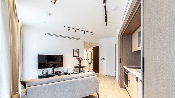 A modern living room with a sofa, a flat-screen TV, a small round table with a vase, and a kitchen area with cabinets and a dishwasher. - Elegant Flat in Notting Hill's Heartbeat (London)