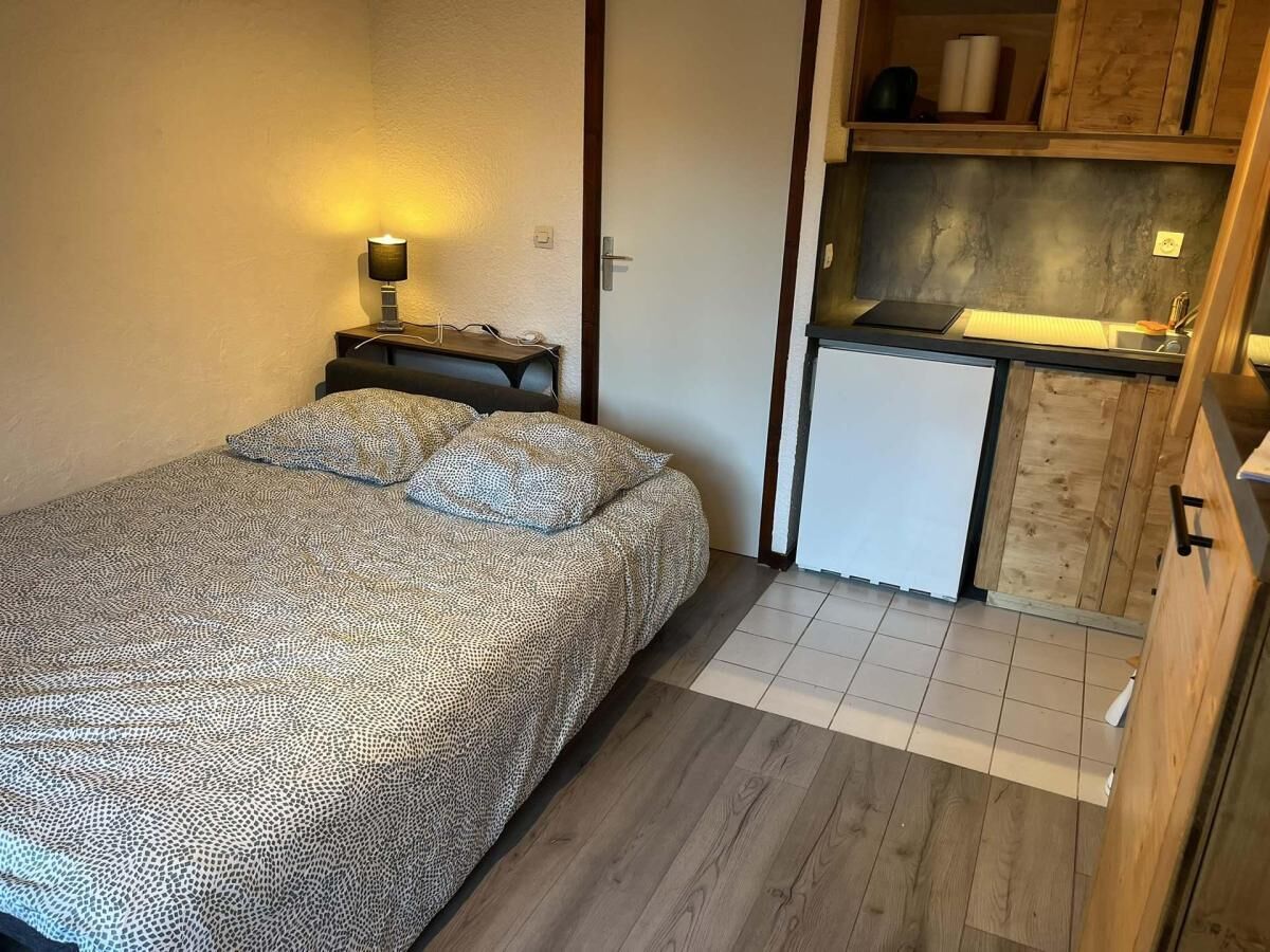 A bedroom with a bed, bedside table, lamp, and a small kitchenette with a sink and cabinets.