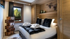 Une chambre moderne avec un plafond en bois, un grand lit recouvert de draps blancs, un plaid en fourrure et une fenêtre donnant sur la forêt.