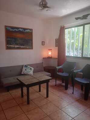 Een woonkamer met een bank, een tafel, twee stoelen, een schilderij, een lamp en een raam met gordijnen.