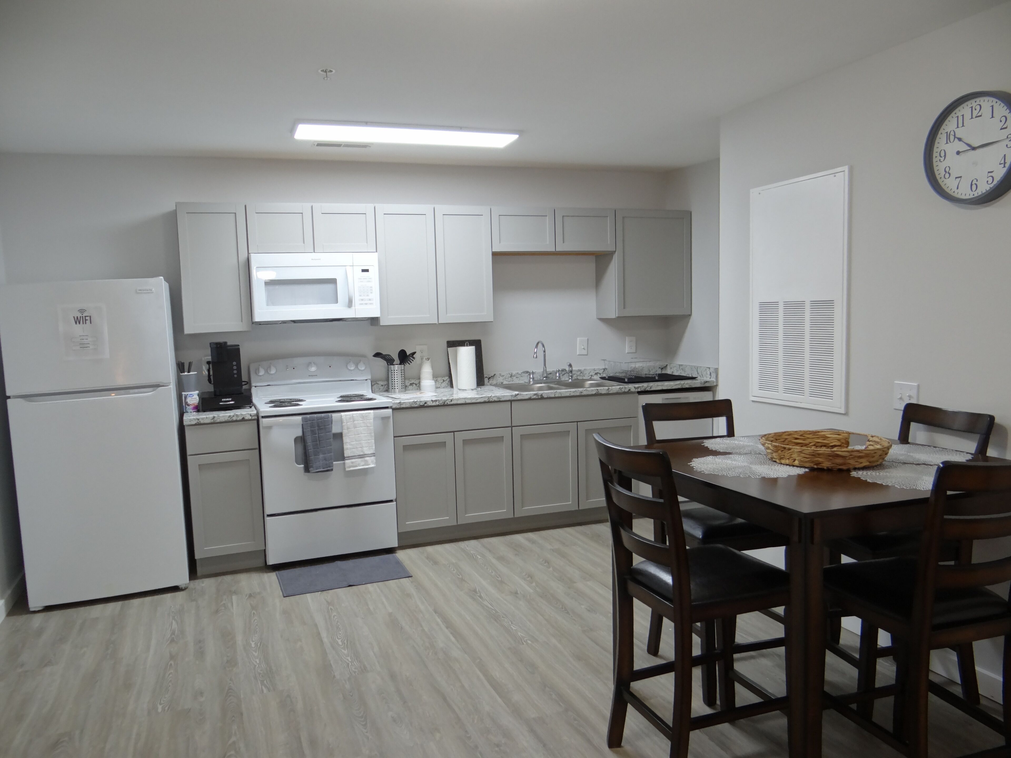 Een kleine keuken met witte apparaten, een eettafel met stoelen en een wandklok.