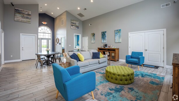 Un ampio soggiorno con un divano grigio, poltrone blu e un poggiapiedi verde. C'è una zona pranzo con tavolo e sedie, e un angolo cottura con un mobiletto in legno. La stanza presenta un soffitto alto con illuminazione a incasso e una grande finestra.