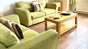 Una sala de estar con dos sofás verdes, una mesa de centro de madera, una lámpara y cuadros enmarcados en la pared.